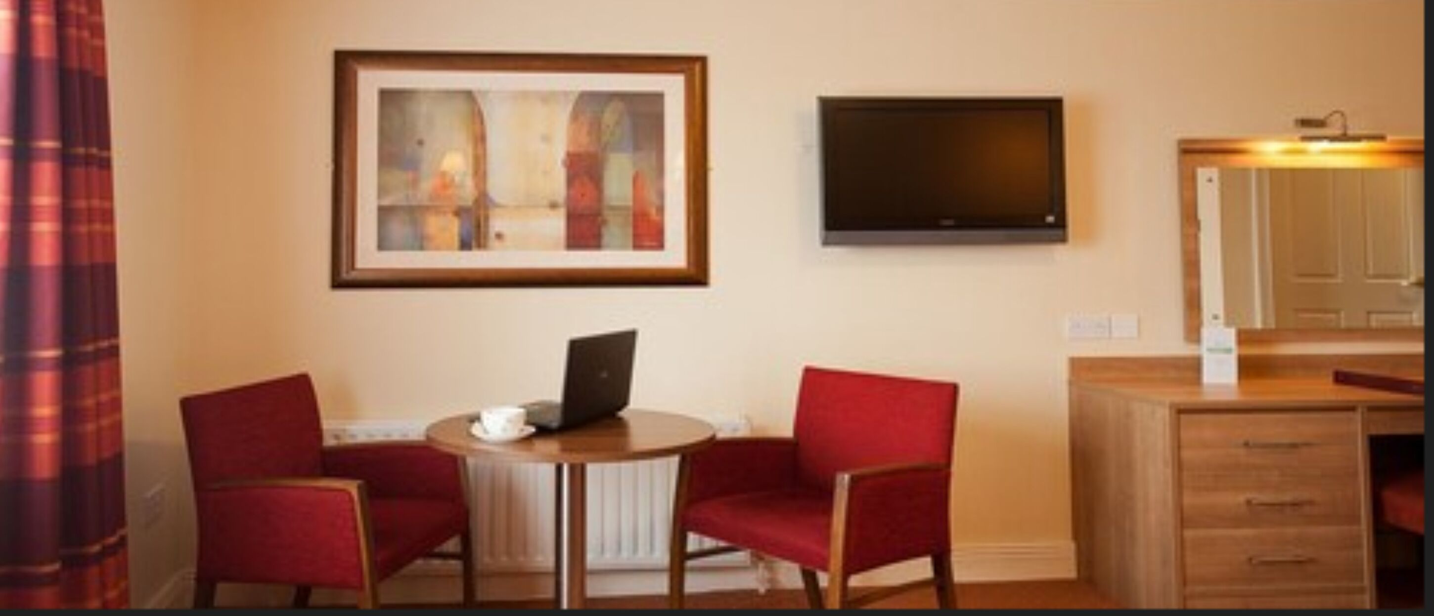 A hotel room with a round table, two red chairs, a laptop, a framed abstract painting, a flat-screen TV, and a wooden dresser.