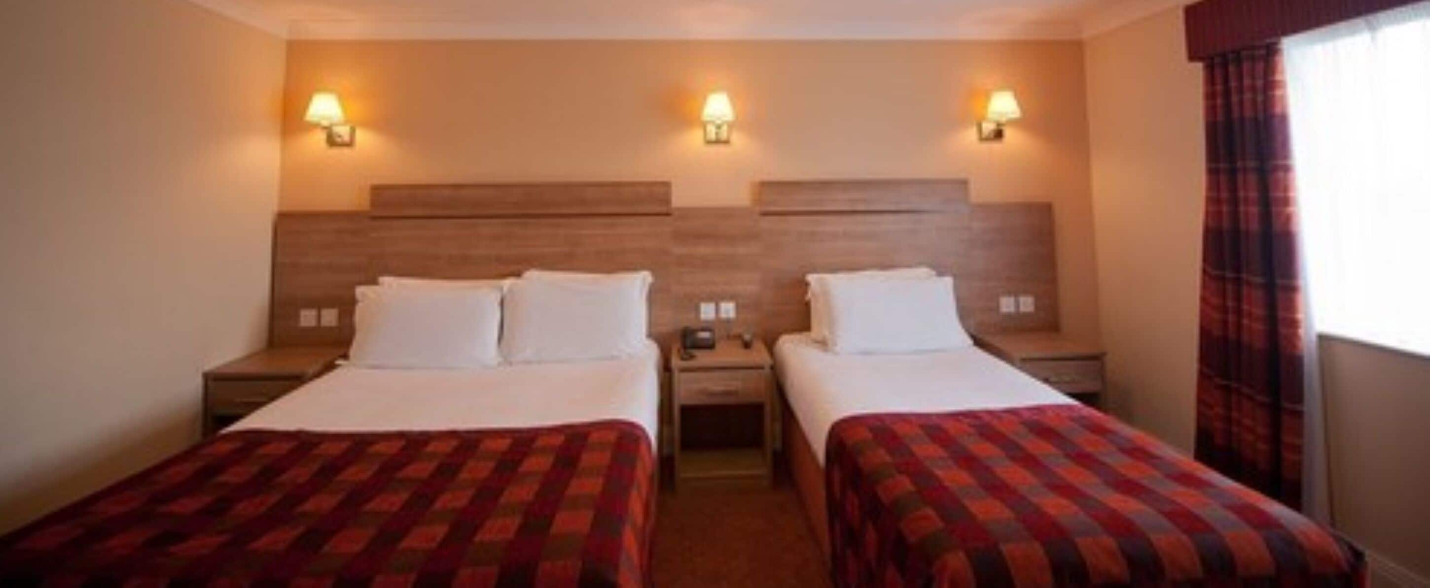 A hotel room with two beds, each with a red and black checkered bedspread, and two bedside tables. There are wall-mounted lamps on either side of the headboard.