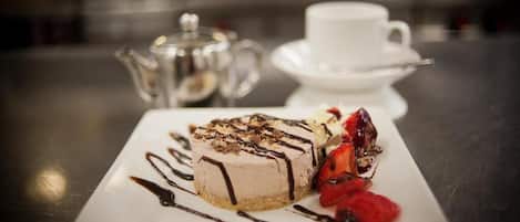 A dessert on a white plate with chocolate drizzle and strawberries, a white cup and saucer, and a teapot on a table.