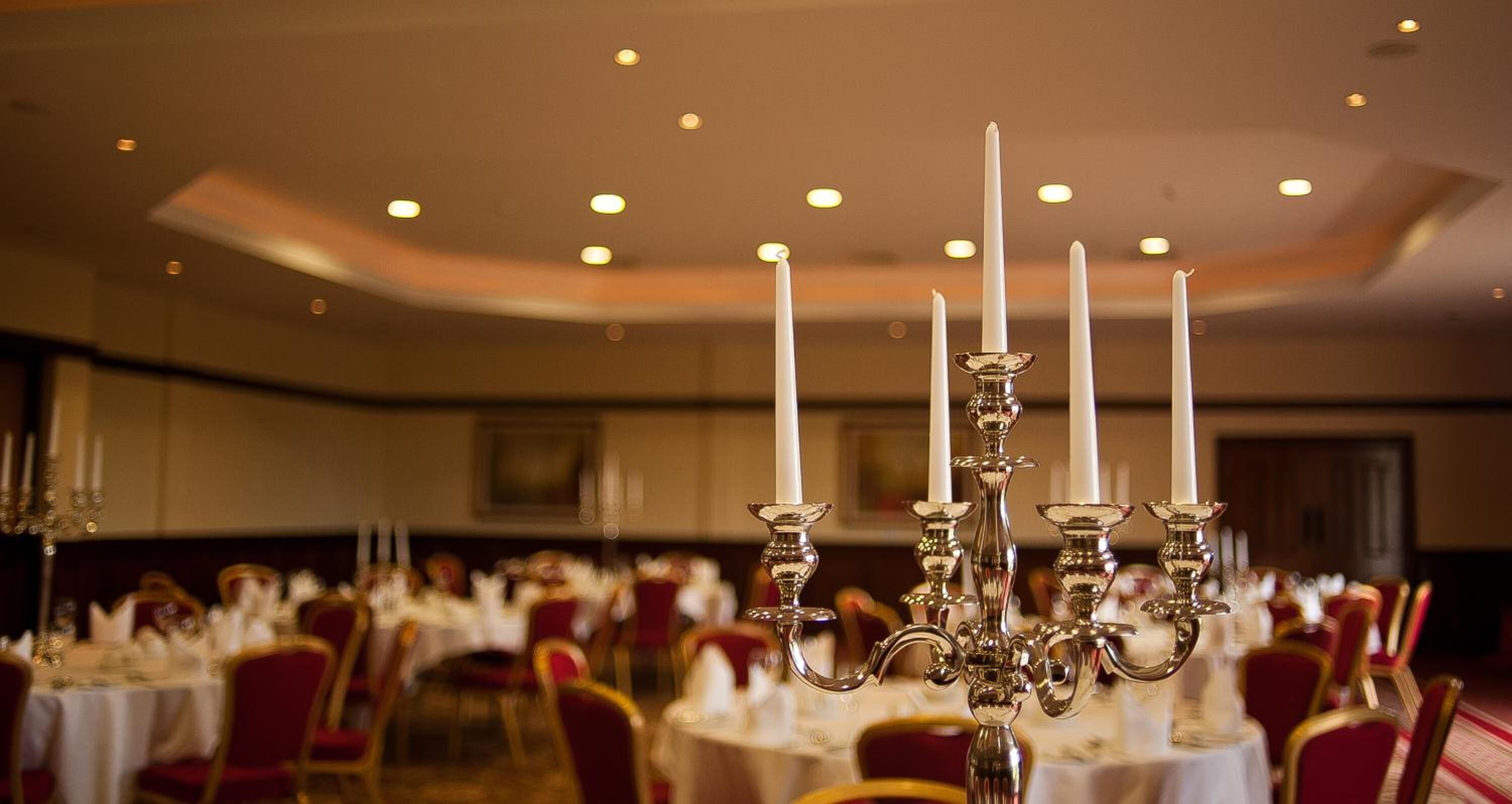 A banquet hall with round tables set for dining, a chandelier with multiple candles, and red upholstered chairs.