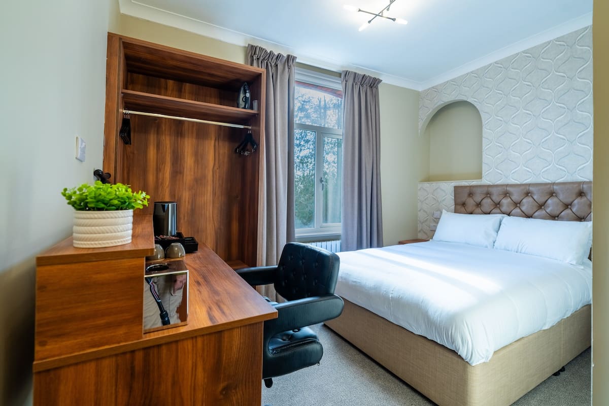 A hotel room with a wooden desk, a comfortable chair, a bed with a tufted headboard, and a window with curtains.