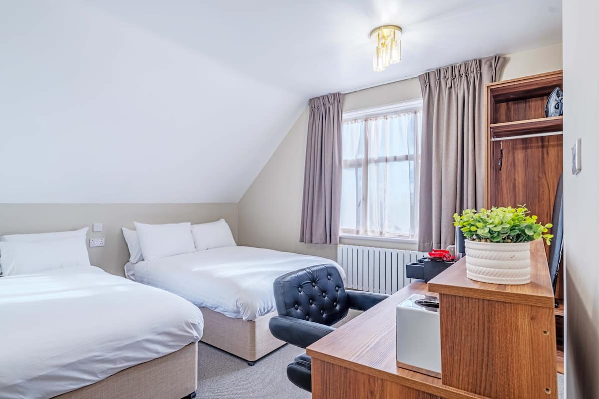 A hotel room with two beds, a desk, a chair, a radiator, a window with curtains, and a ceiling light fixture.