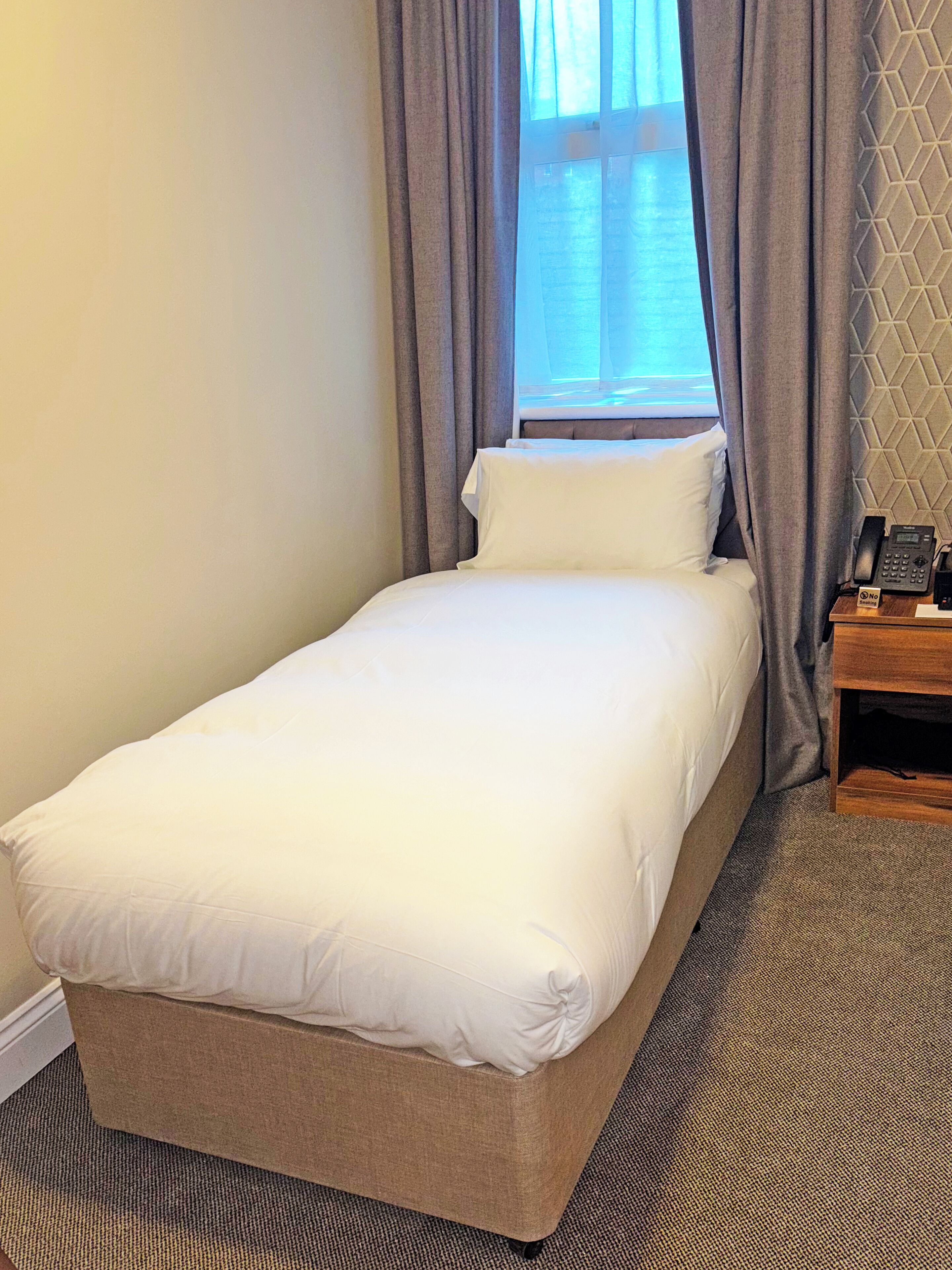 A single bed with white bedding, a wooden nightstand with a telephone, and a window with curtains.