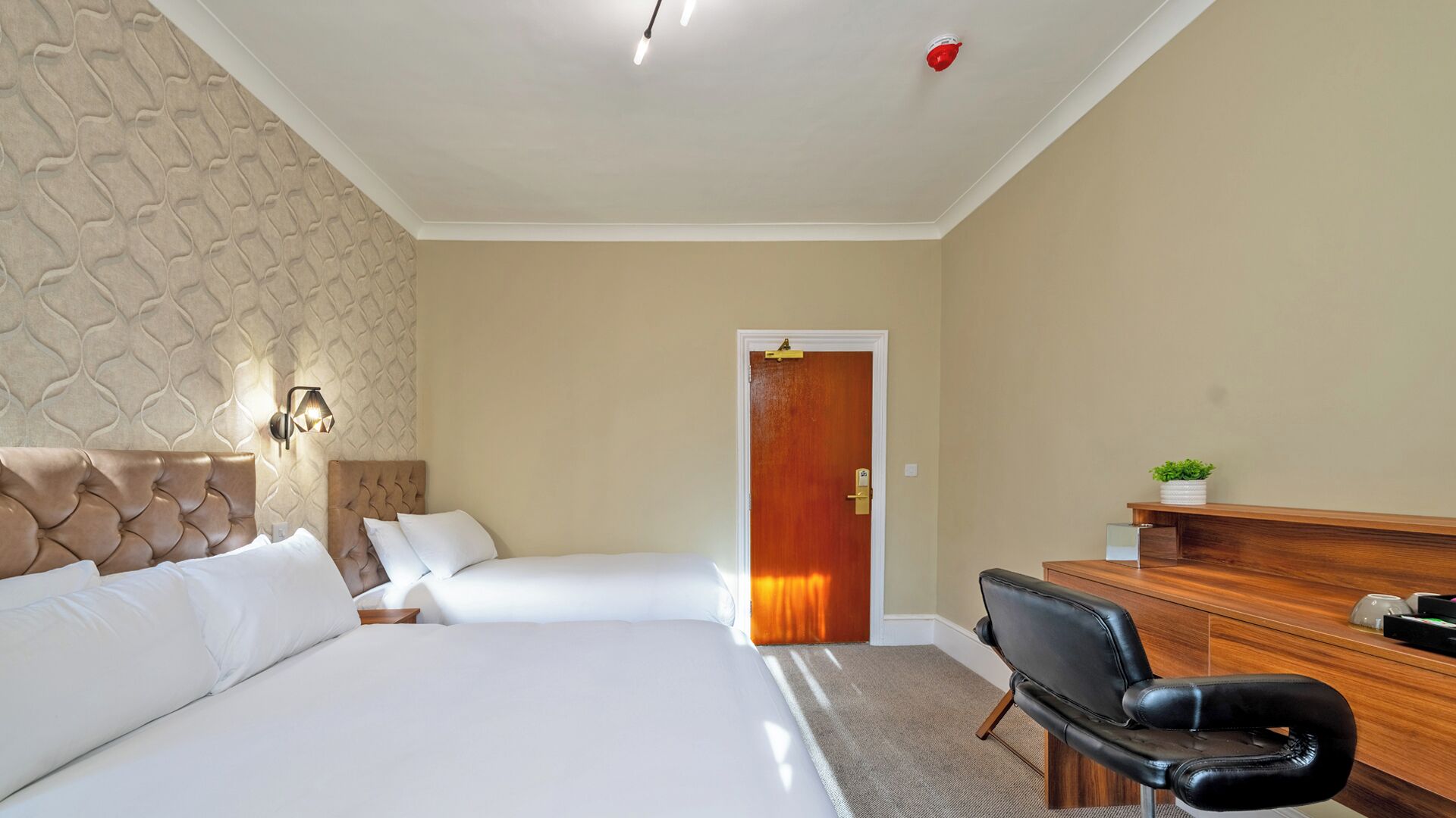 Une chambre d’hôtel avec deux lits, un bureau, une chaise, une porte en bois et un mur orné d’un motif texturé.