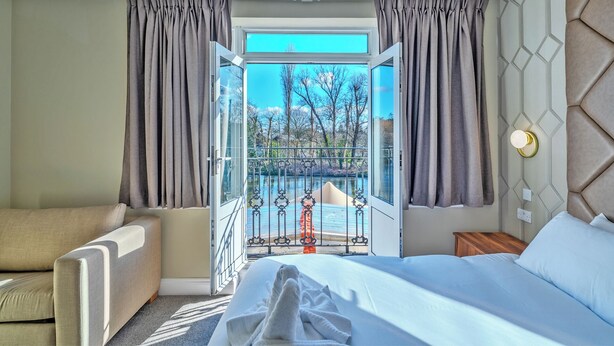 A hotel room with a bed, a sofa, a view of a body of water, and a balcony with a railing. - Uno Thames Hotel (Maidenhead)