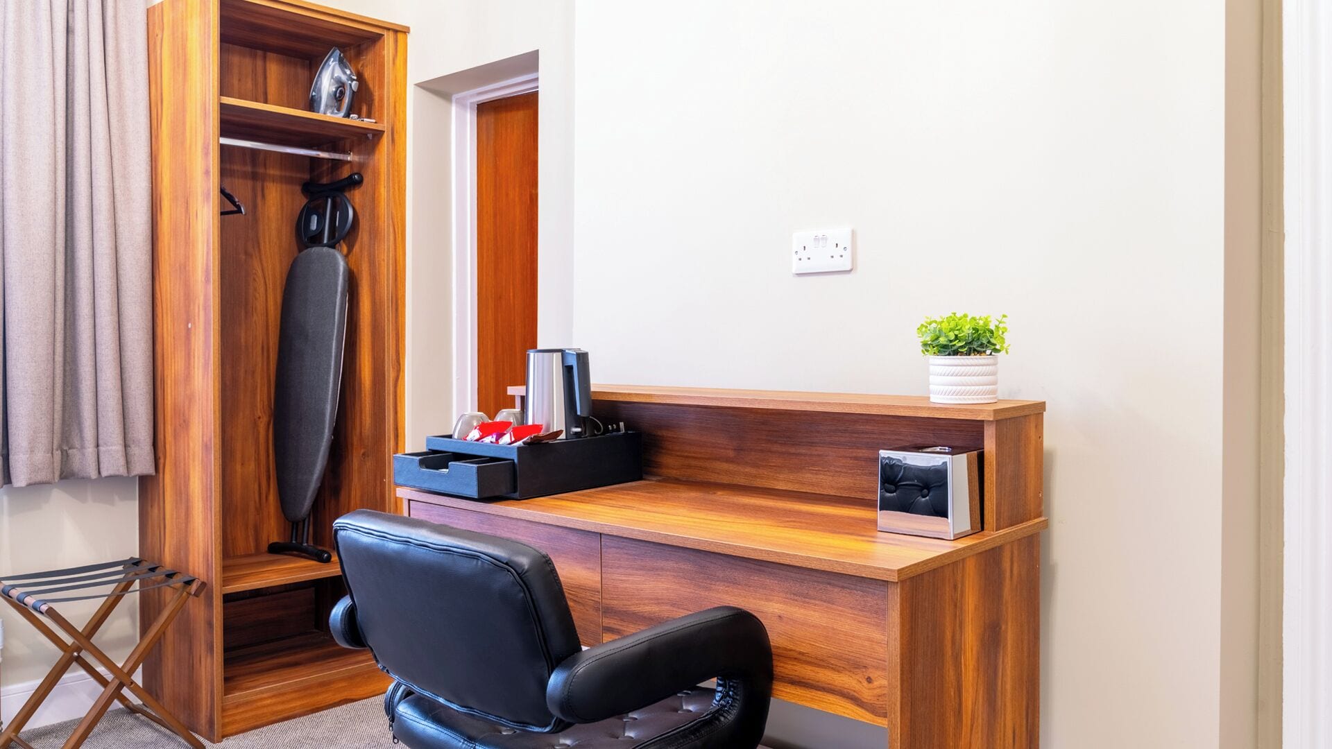 A compact office space with a wooden desk, a black office chair, a shelf with a kettle, and a small potted plant.