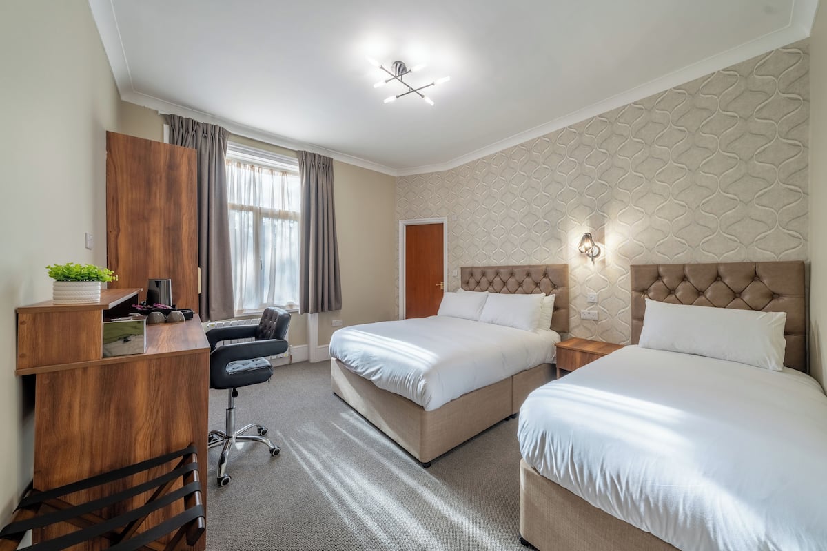A hotel room with a desk, two beds, a chair, and a window with curtains.