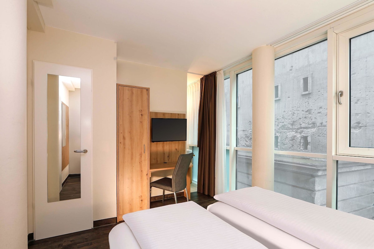 A hotel room with a bed, a desk, a chair, a TV, and large windows.