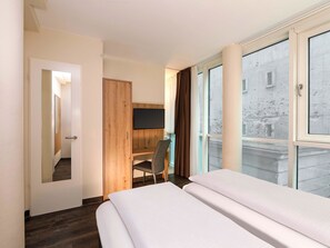 A hotel room with a bed, a desk, a chair, a TV, and large windows. - B&B HOTEL Berlin-Mitte (Berlin)