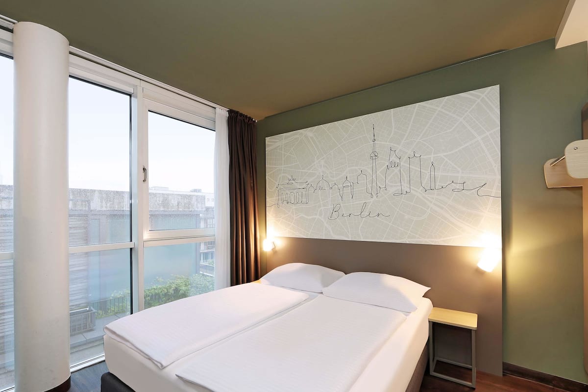 A hotel room with a large bed, a cityscape wall art, a window with curtains, and a nightstand.