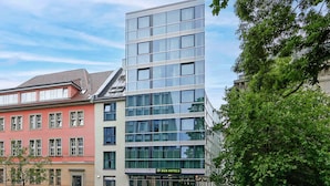 Un hotel moderno con fachada de vidrio, un edificio rojo contiguo y una calle bordeada de árboles.