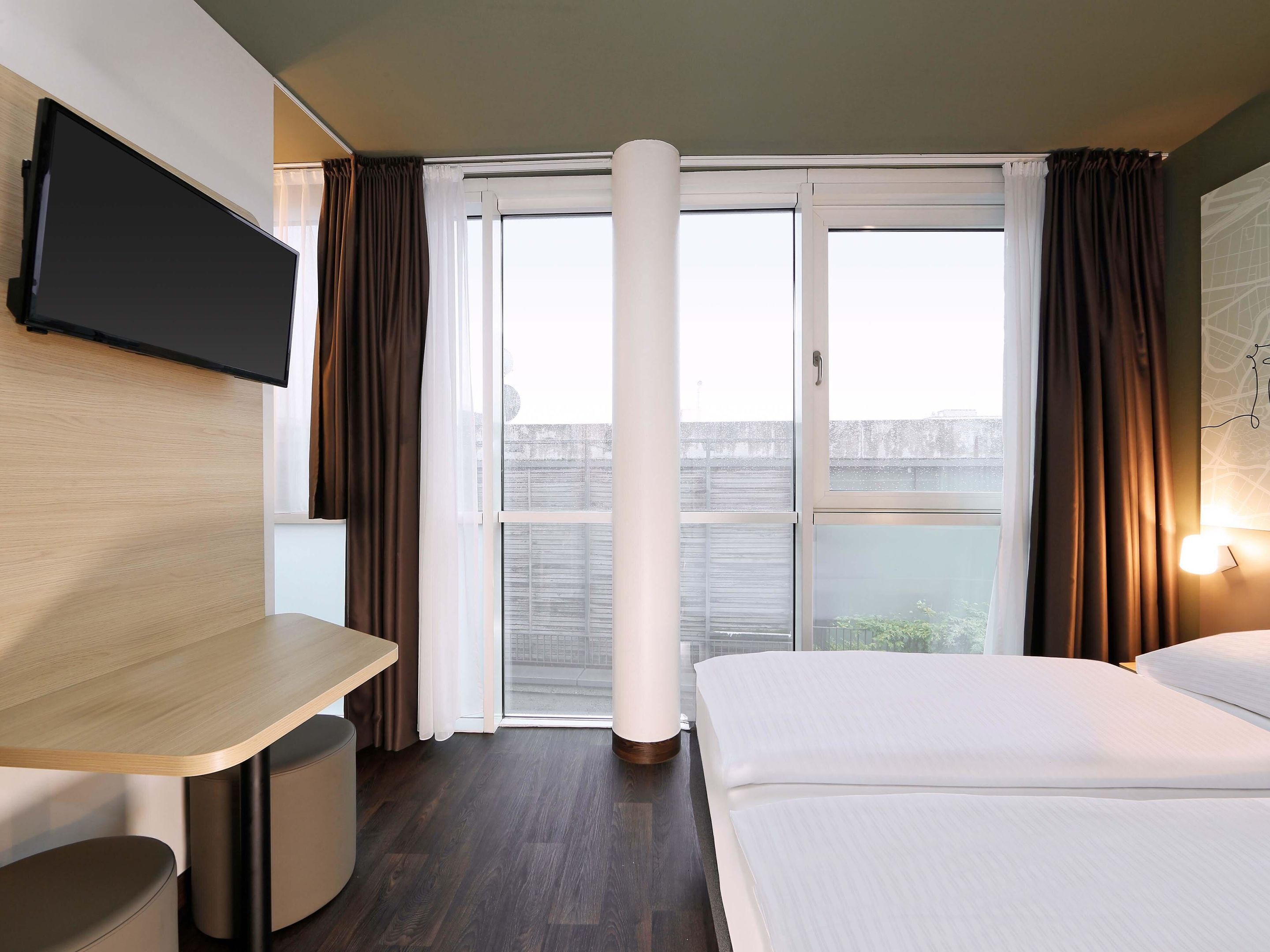 A hotel room with a bed, a desk, a TV mounted on the wall, and a large window with curtains.