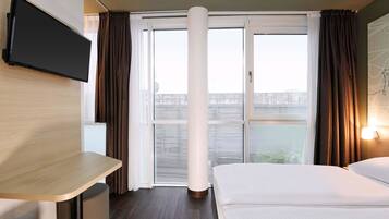 A hotel room with a bed, a desk, a TV mounted on the wall, and a large window with curtains.