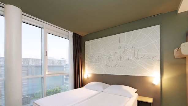 Una habitación de hotel con una cama grande, un cuadro con temática urbana, una ventana con cortinas y una mesita de noche.