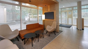 Un área de espera moderna con techo de listones de madera, un sofá, dos sillones, un televisor y amplias ventanas con vistas al exterior.