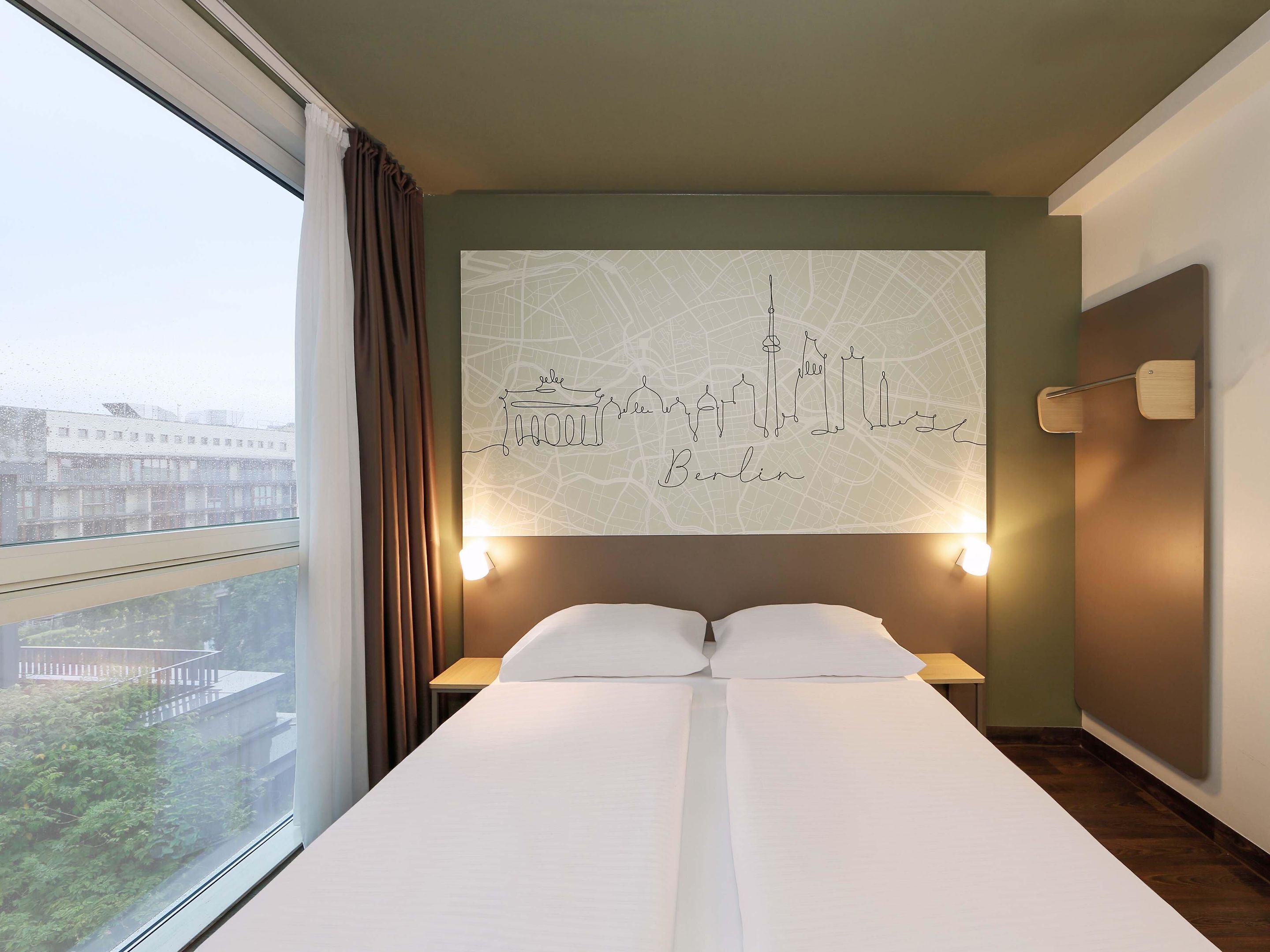 A hotel room with a large bed, a cityscape wall art, a window with curtains, and a nightstand with a lamp.