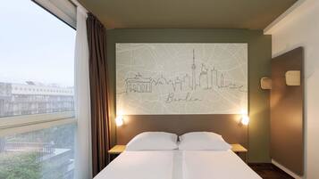 A hotel room with a large bed, a cityscape wall art, a window with curtains, and a nightstand with a lamp.