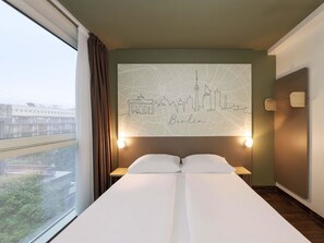 A hotel room with a large bed, a cityscape wall art, a window with curtains, and a nightstand with a lamp. - B&B HOTEL Berlin-Mitte (Berlin)