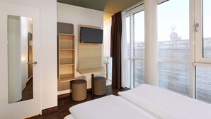 Habitación de hotel con cama, televisor montado en la pared, ventana con cortinas y vista a la ciudad.