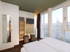 A hotel room with a bed, a TV mounted on the wall, a window with curtains, and a view of the city. - B&B HOTEL Berlin-Mitte (Berlin)