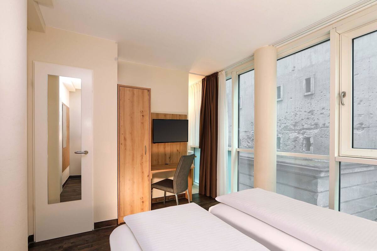 A hotel room with a bed, a desk, a chair, a TV, and large windows.