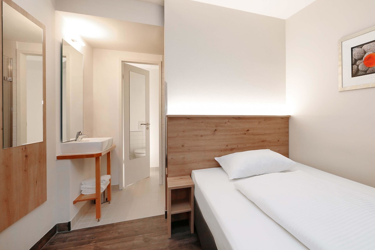 A hotel room with a wooden headboard, a bed with white bedding, a bedside table, a mirror, and a bathroom with a sink and toilet.