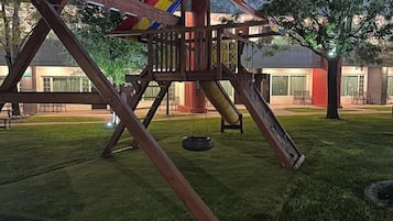 A playground with a swing set and a slide, surrounded by grass and trees.
