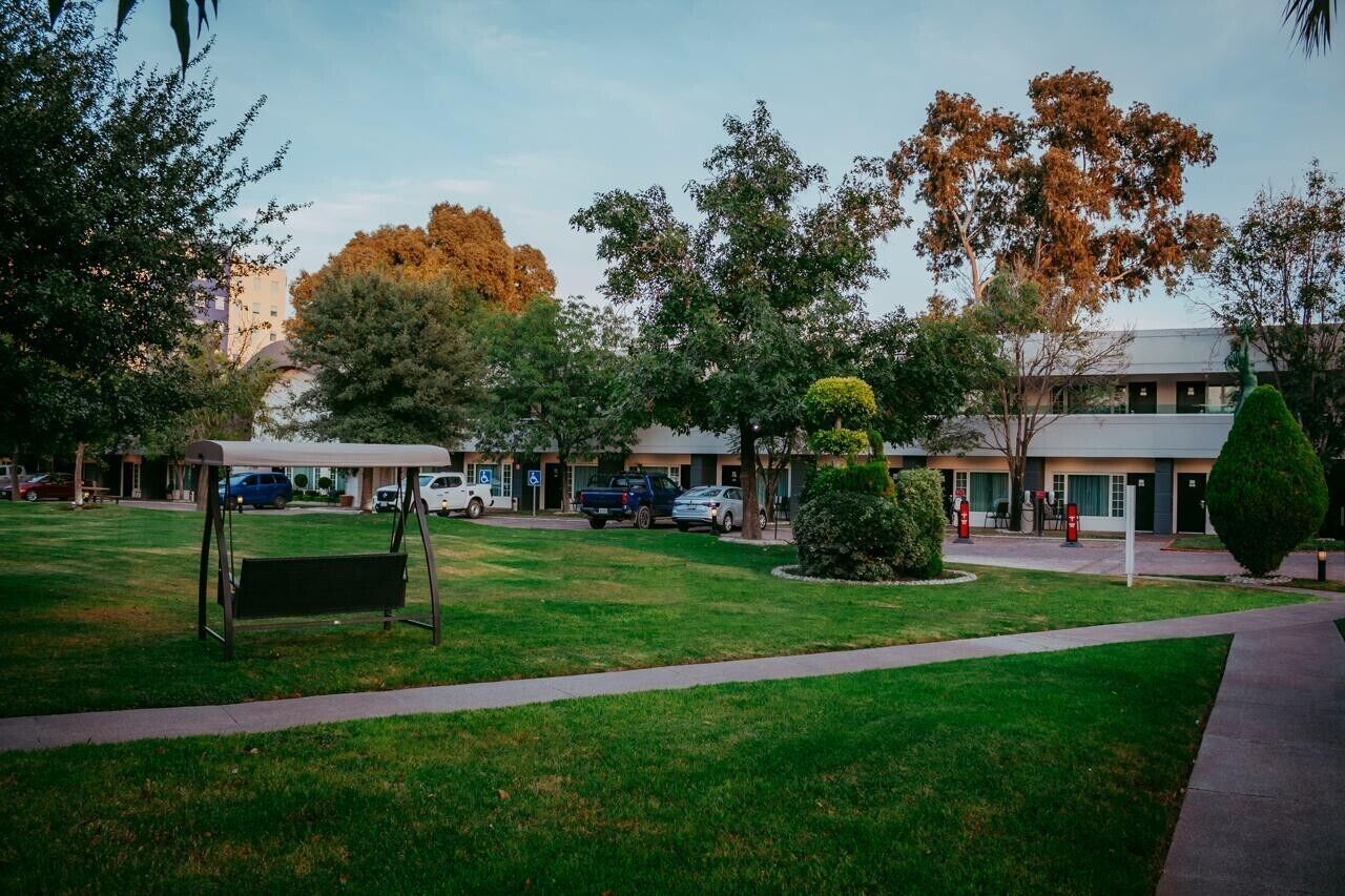 A grassy area with a bench, trees, and a building with a parking lot in the background.
