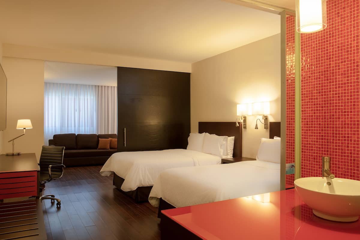 Ein Hotelzimmer mit zwei Betten, einem Schreibtisch, einem Stuhl, einer Schreibtischlampe, einem Nachttisch, einem Spiegel und einer Wand mit roten Fliesen.