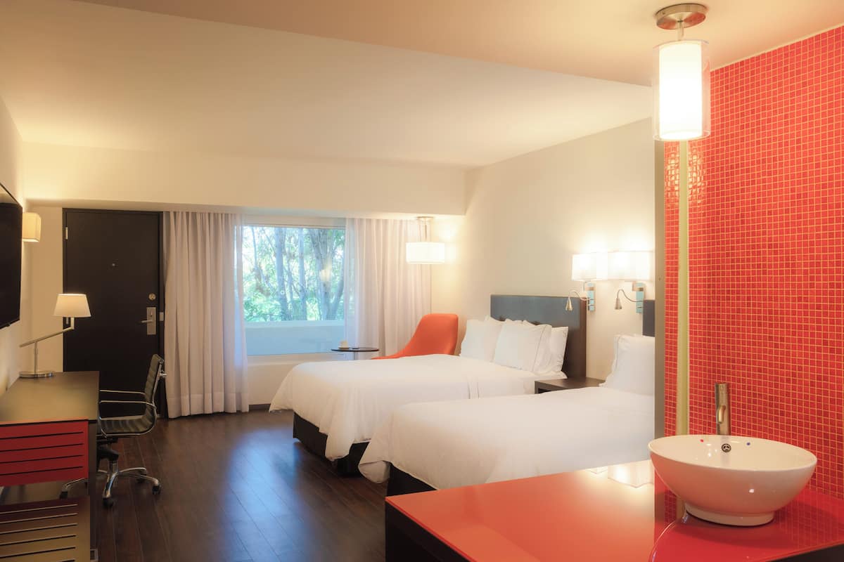 Ein Hotelzimmer mit zwei Betten, einem Schreibtisch, einem Stuhl, einer roten Akzentwand und einem großen Fenster.