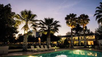 A pool area with lounge chairs, palm trees, and a hotel building in the background.