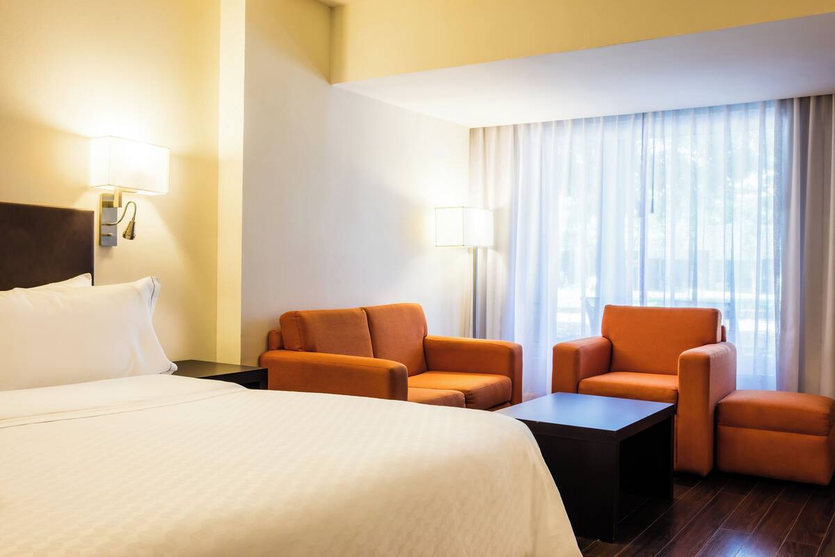 Ein Hotelzimmer mit einem großen Bett, zwei orangefarbenen Sesseln, einem kleinen Tisch und einem Fenster mit transparenten Vorhängen.