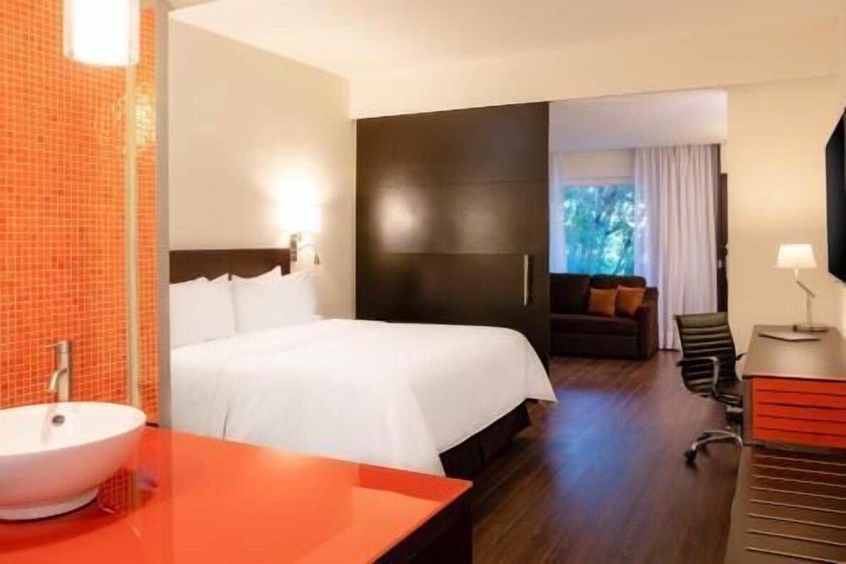 A hotel room with a bed, a sink, a dark-colored wardrobe, a small seating area with a sofa and chairs, and a window with a view of trees.