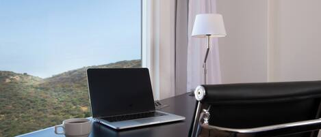 Una computadora portátil sobre un escritorio con una taza, una lámpara y una silla, con vista a un paisaje montañoso.