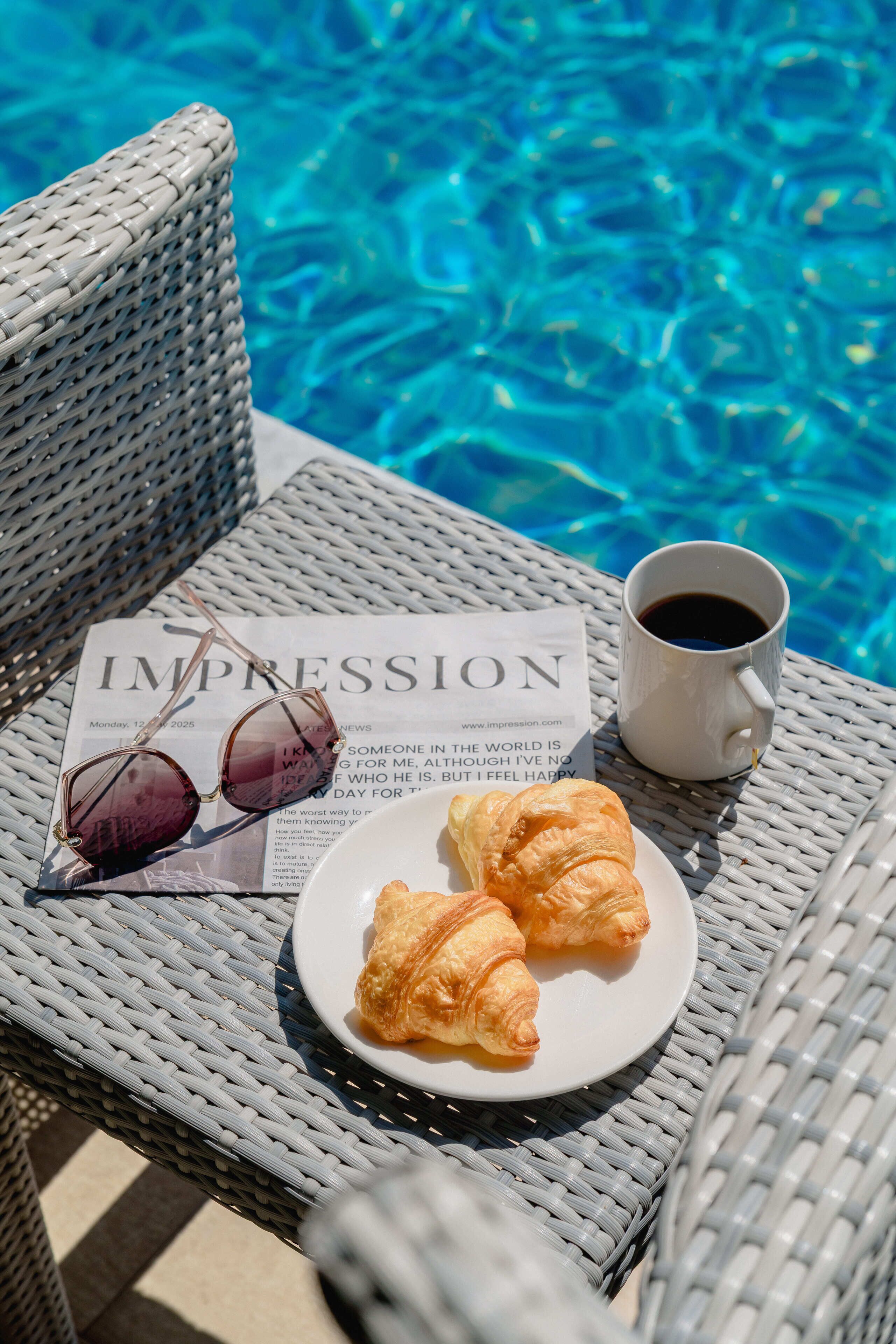 Una silla de mimbre junto a una piscina con un plato de croissants, una taza de café y gafas de sol sobre una revista.