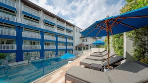 Un área de piscina en la azotea con sillones de descanso y sombrillas, un cielo azul despejado y un edificio con paredes exteriores azules y blancas.