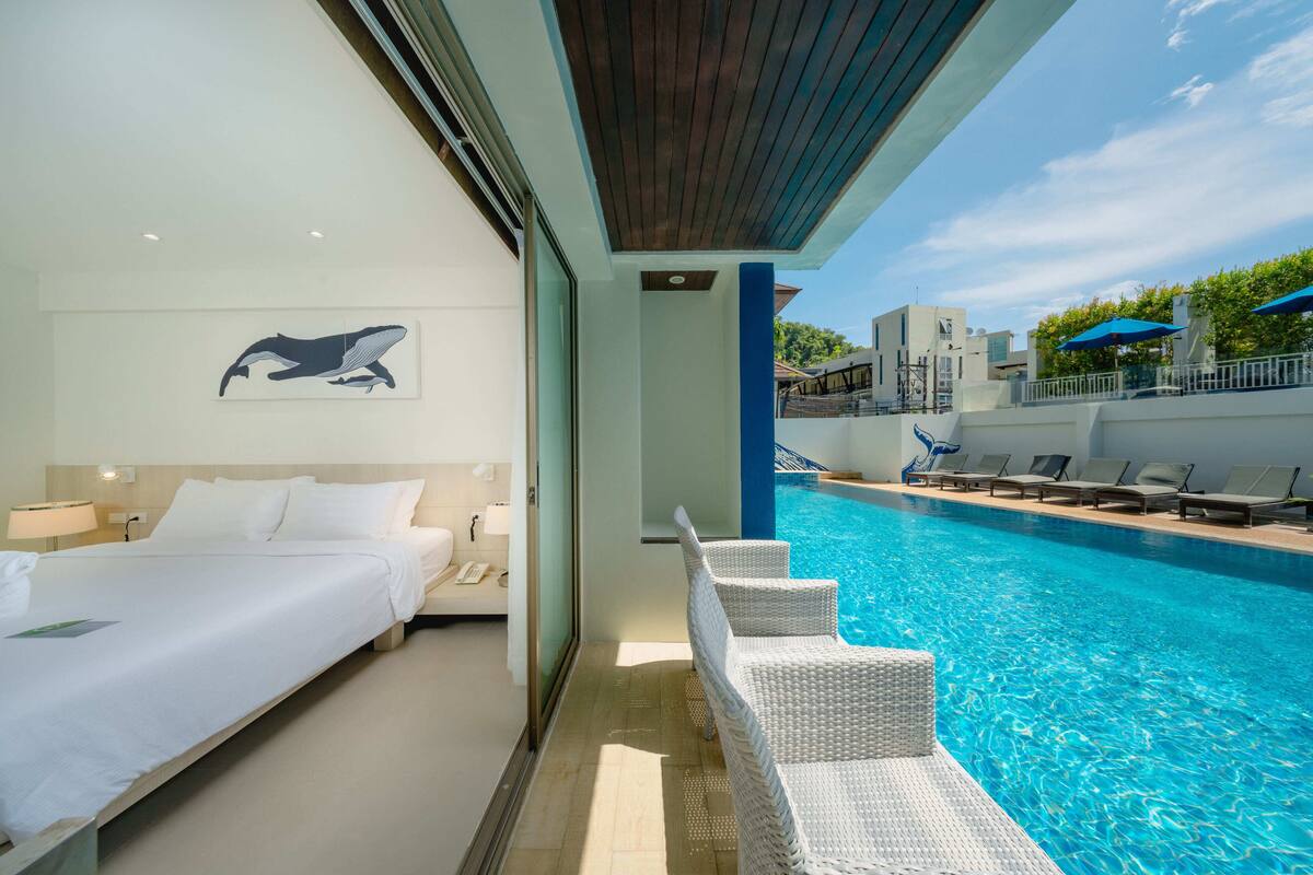 Habitación de hotel con cama, un ventanal grande y vistas a una piscina con sillas de descanso.