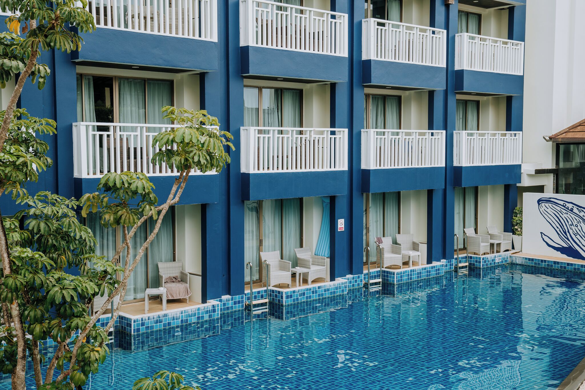 A hotel with a pool area, featuring multiple balconies with white railings, a tiled pool, and lounge chairs.