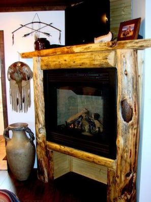 A rustic wooden fireplace with a stone surround, a decorative wall hanging, and a vase on the floor.