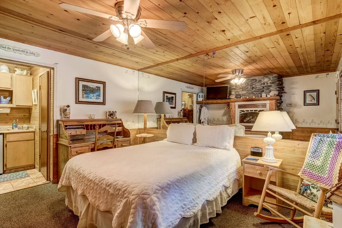 A cozy bedroom with a large bed, wooden ceiling, and a stone fireplace.