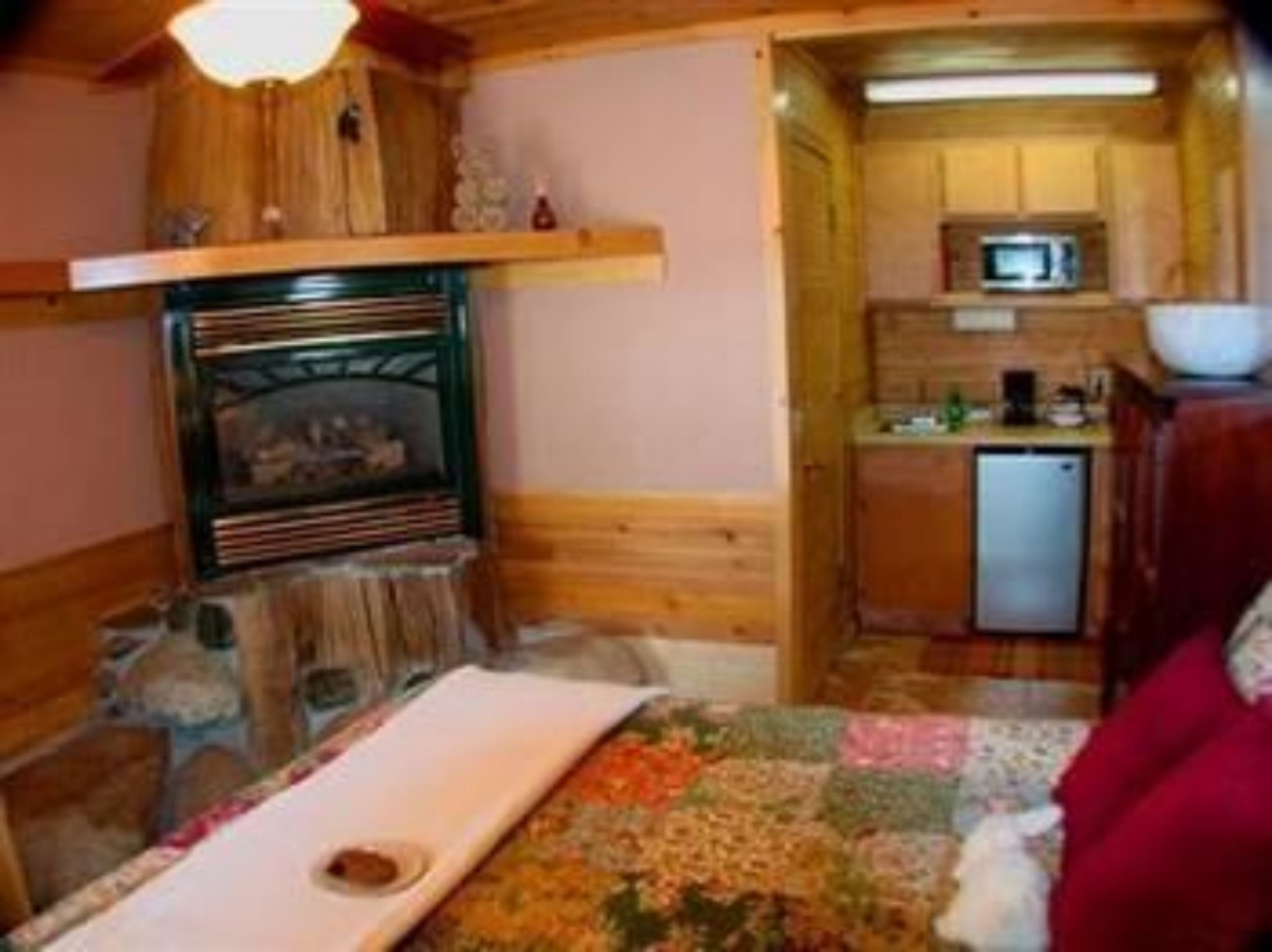 A compact wooden cabin interior with a fireplace, a small kitchenette, and a bed with a quilt.