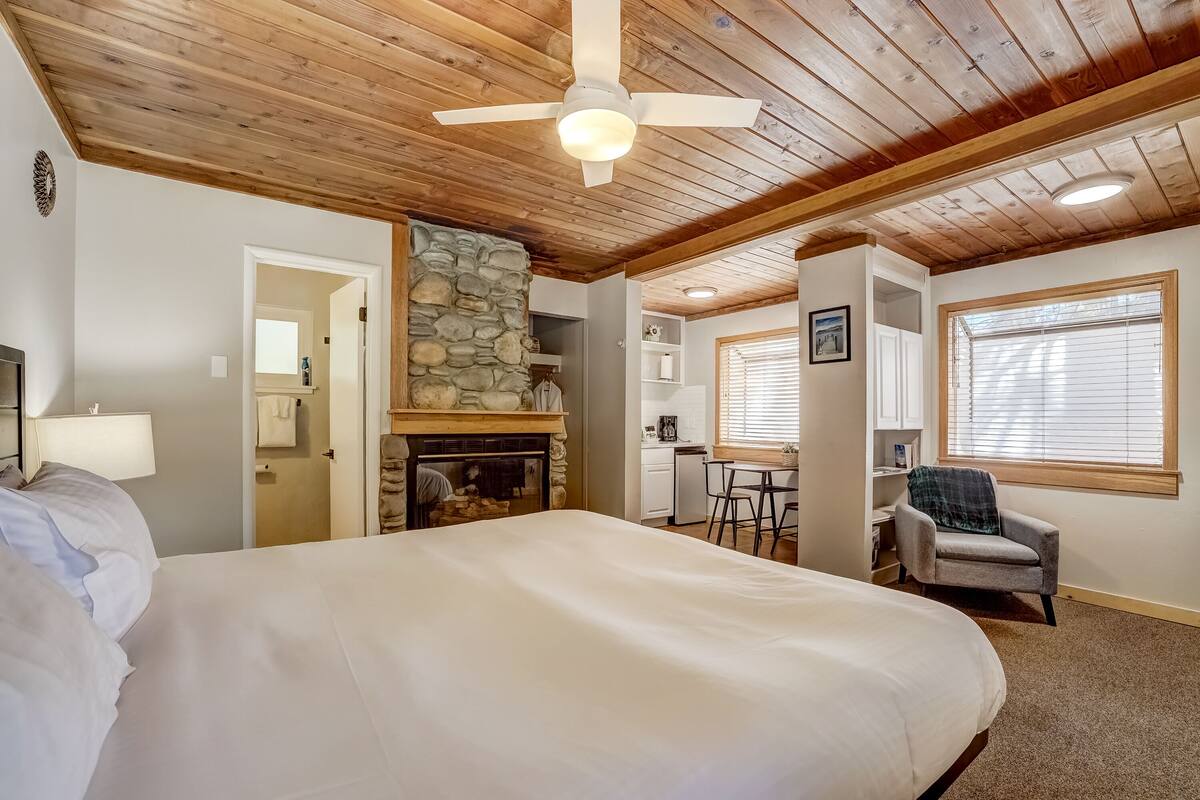 A bedroom with a large bed, a stone fireplace, a ceiling fan, and a view of the kitchen.