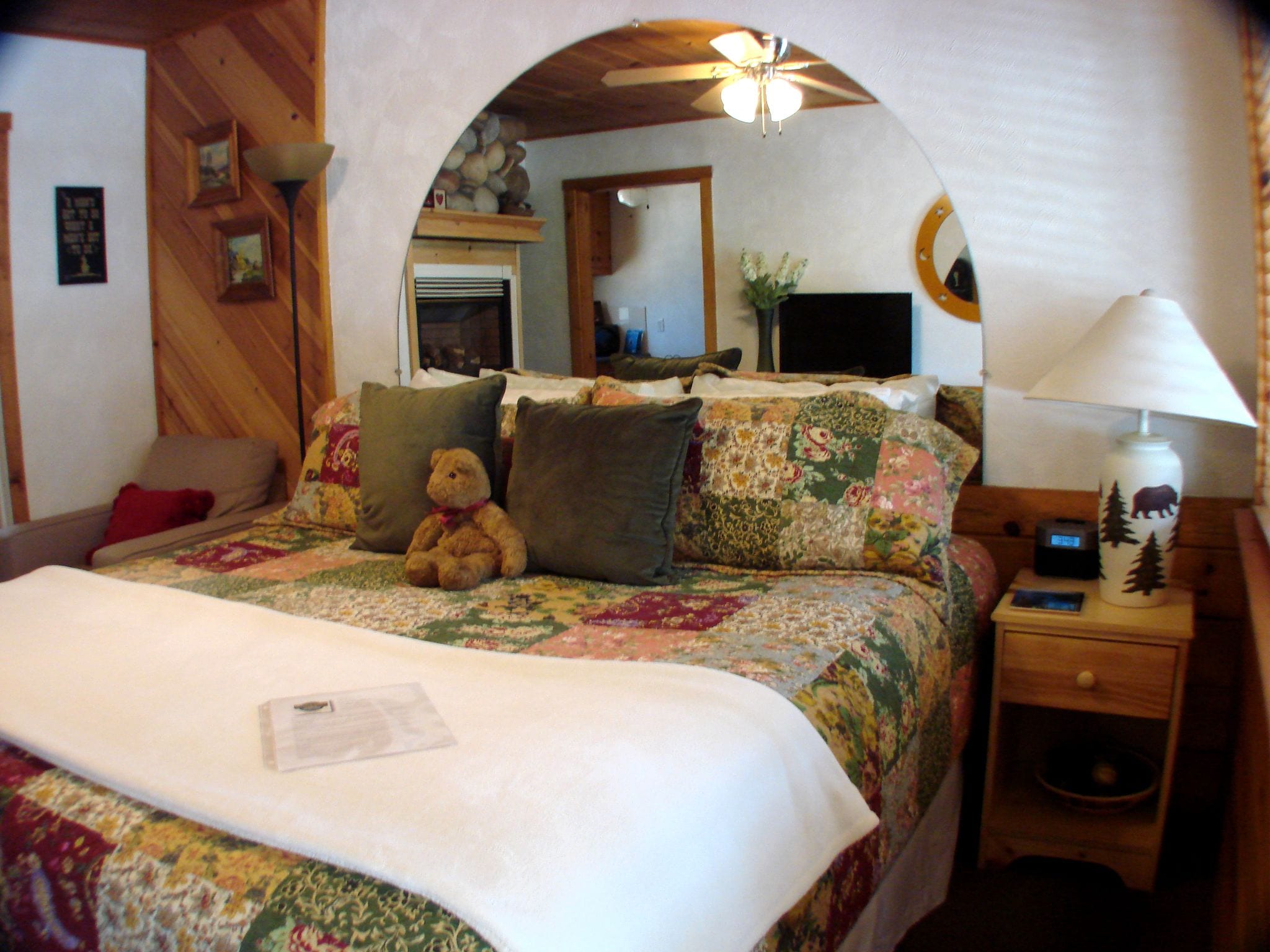 A cozy bedroom with a wooden ceiling, a bed with floral bedding, a teddy bear, and a nightstand with a lamp.