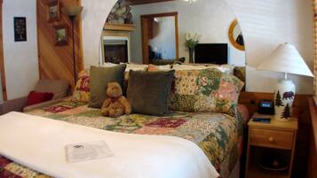 A cozy bedroom with a wooden ceiling, a bed with floral bedding, a teddy bear, and a nightstand with a lamp.