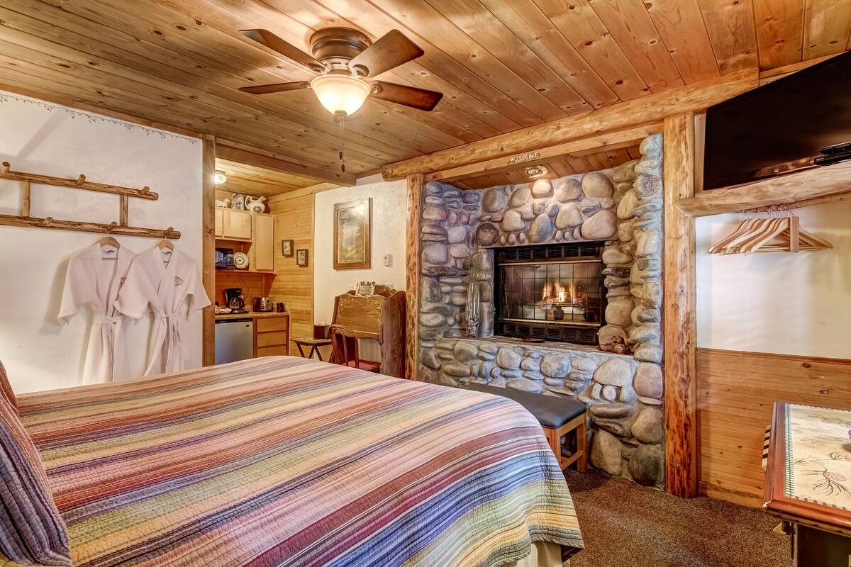 A cozy room with a bed, a stone fireplace, wooden walls, and a ceiling fan.