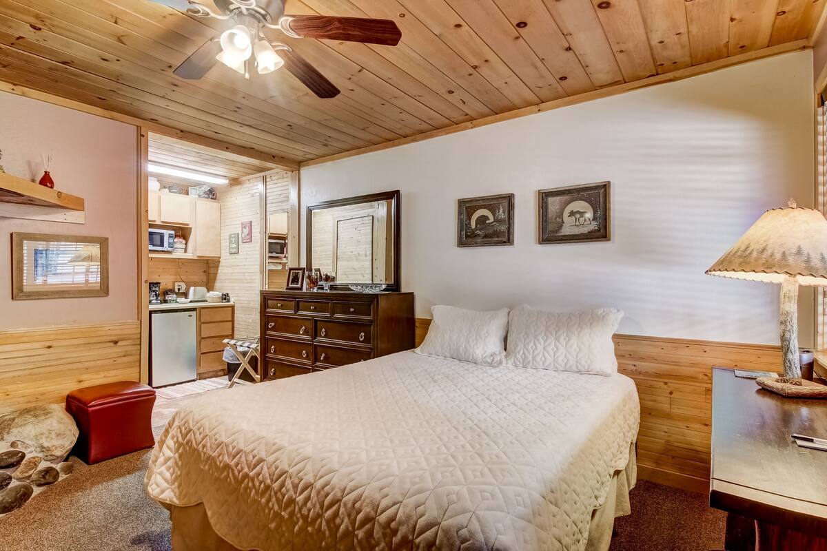 A cozy bedroom with a bed, wooden ceiling, a dresser, a desk, and a lamp.