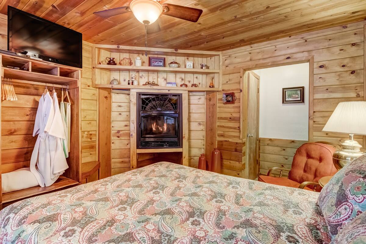A cozy room with a fireplace, wooden walls, a bed with a patterned bedspread, a ceiling fan, and a wooden wardrobe.