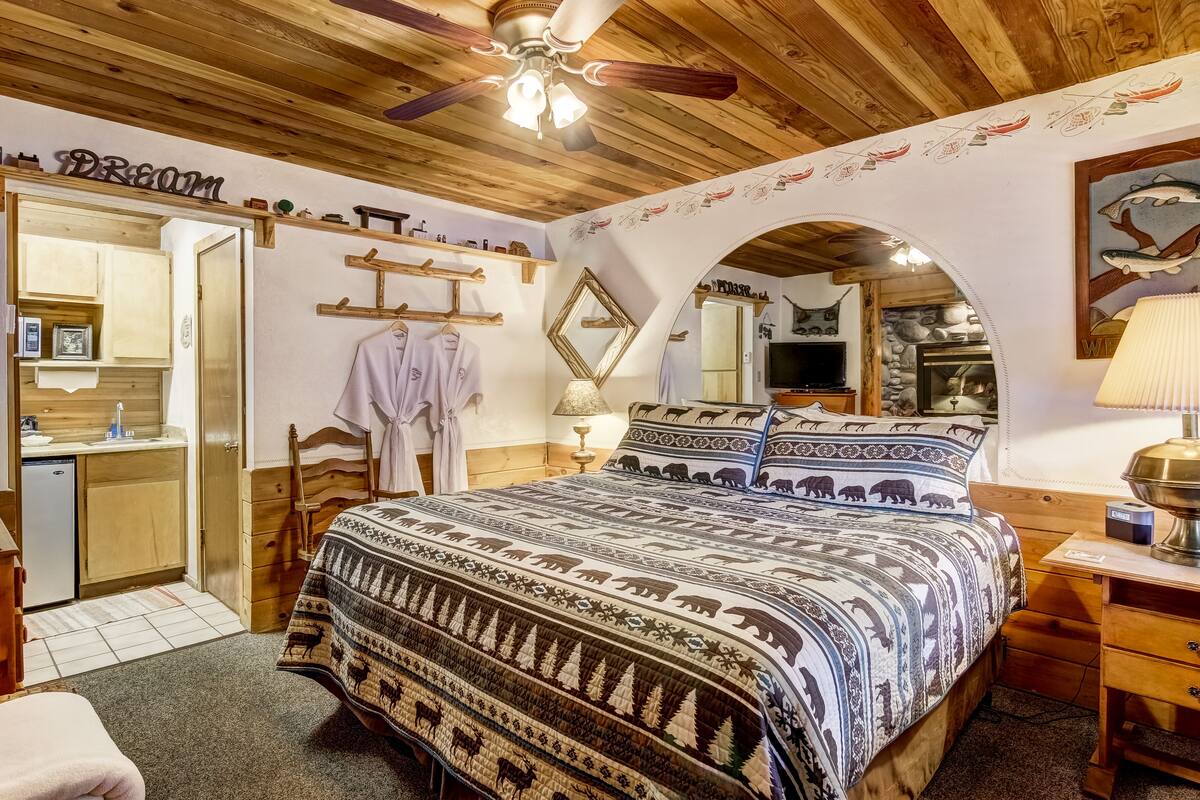 A cozy bedroom with a wooden ceiling, a bed with patterned bedding, a mirror, and a small desk with a lamp.