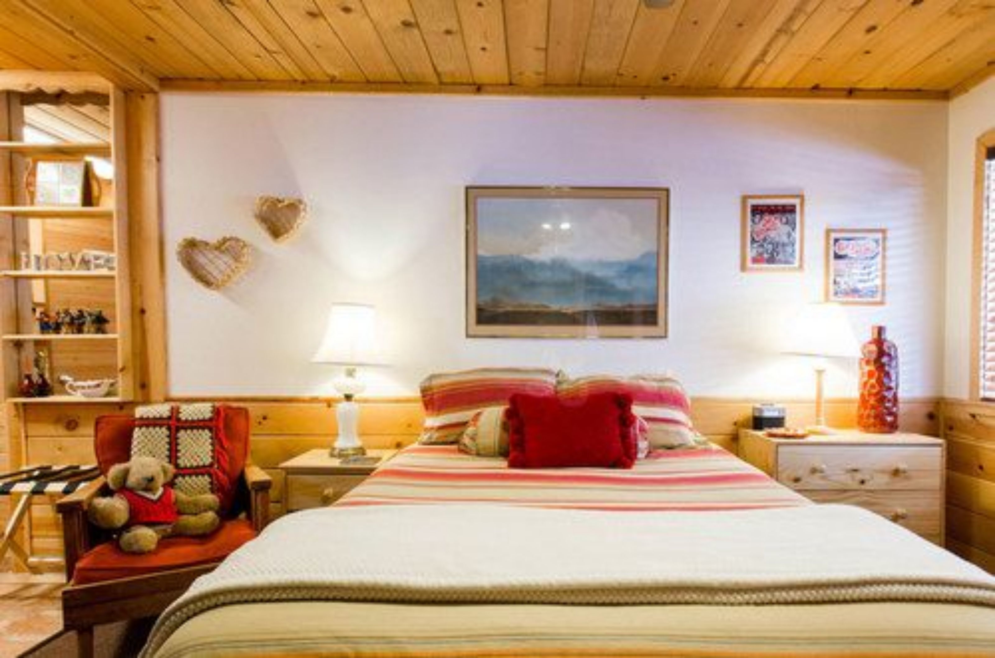 Un dormitorio acogedor con cama, mesita de noche, lámpara, un oso de peluche y estantería con libros.