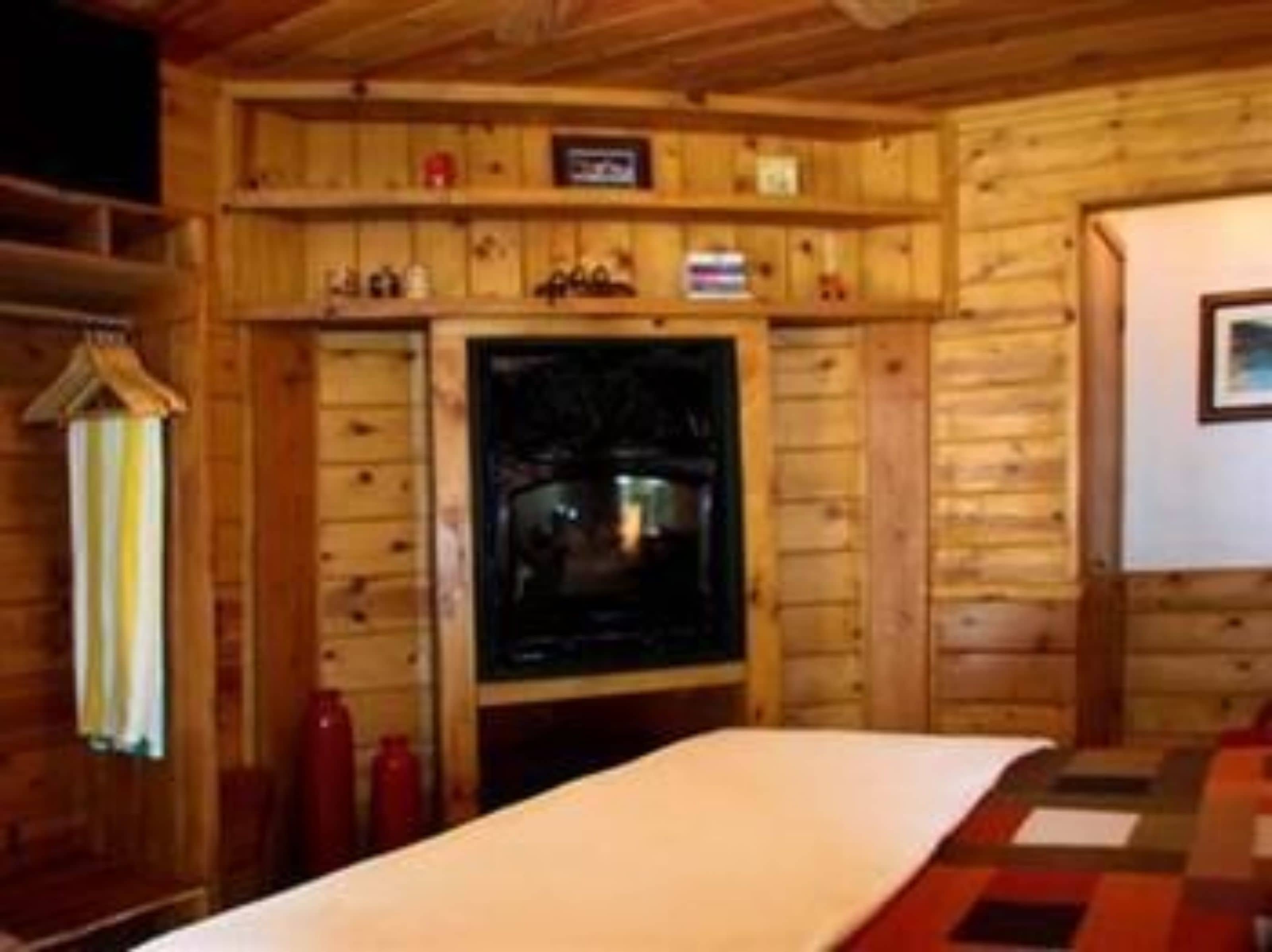 A wooden cabin interior with a bed, a shelf with various items, and a fireplace.